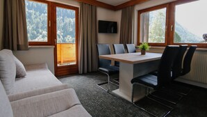 A room with a sofa, a table with chairs, a TV mounted on the wall, and large windows with curtains. - Hotel Tia Apart (Kaunertal)