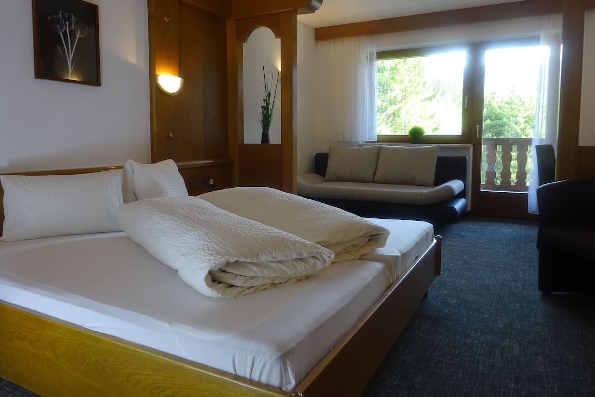 A hotel room with a large bed, a sofa, and a balcony with a view of greenery.