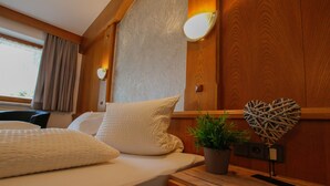 A neatly made bed with white bedding, a potted plant, and a decorative heart-shaped wall hanging in a wooden room. - Hotel Tia Apart (Kaunertal)