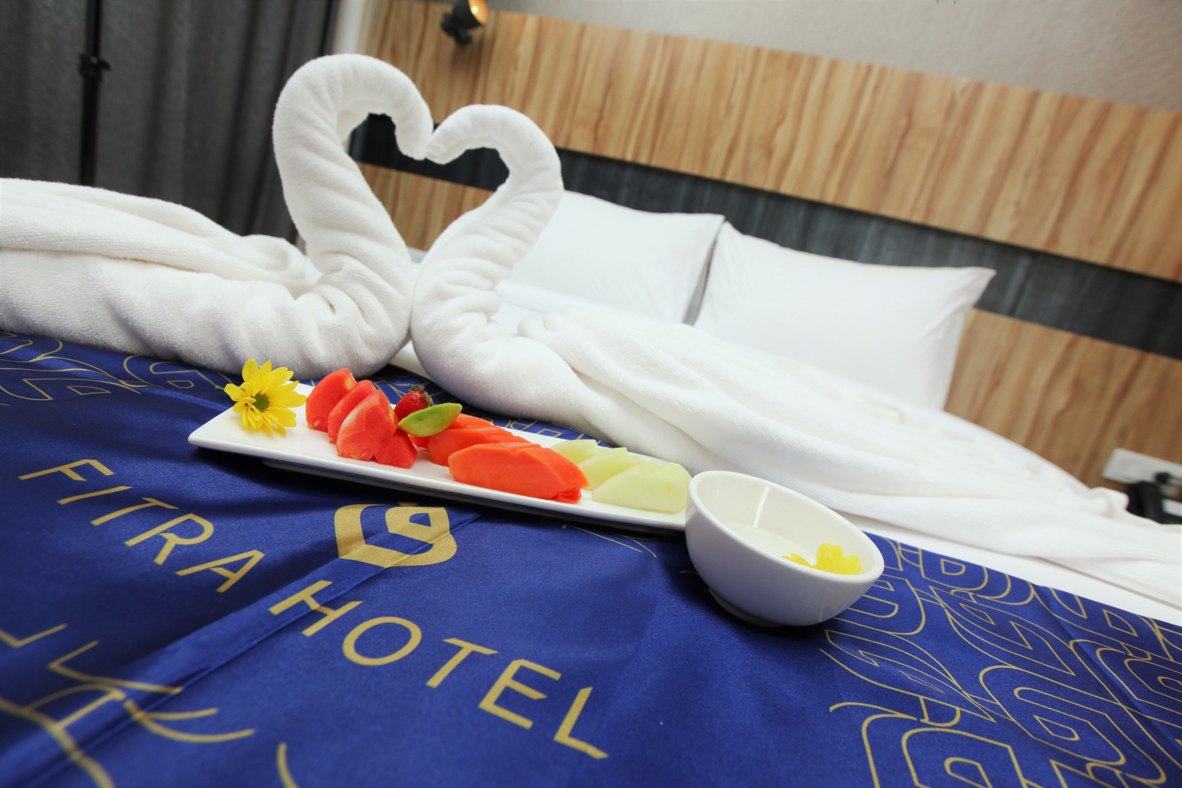 Ein Bett mit einer blauen Hoteldecke, einem Teller Obst, einer Schüssel Joghurt und einer dekorativen Handtuchgestaltung als Schwan.