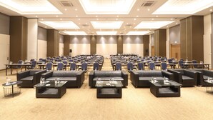 Un amplio salón de conferencias con filas de mesas y sillas, un escenario y una zona de estar con sofás y mesas auxiliares.