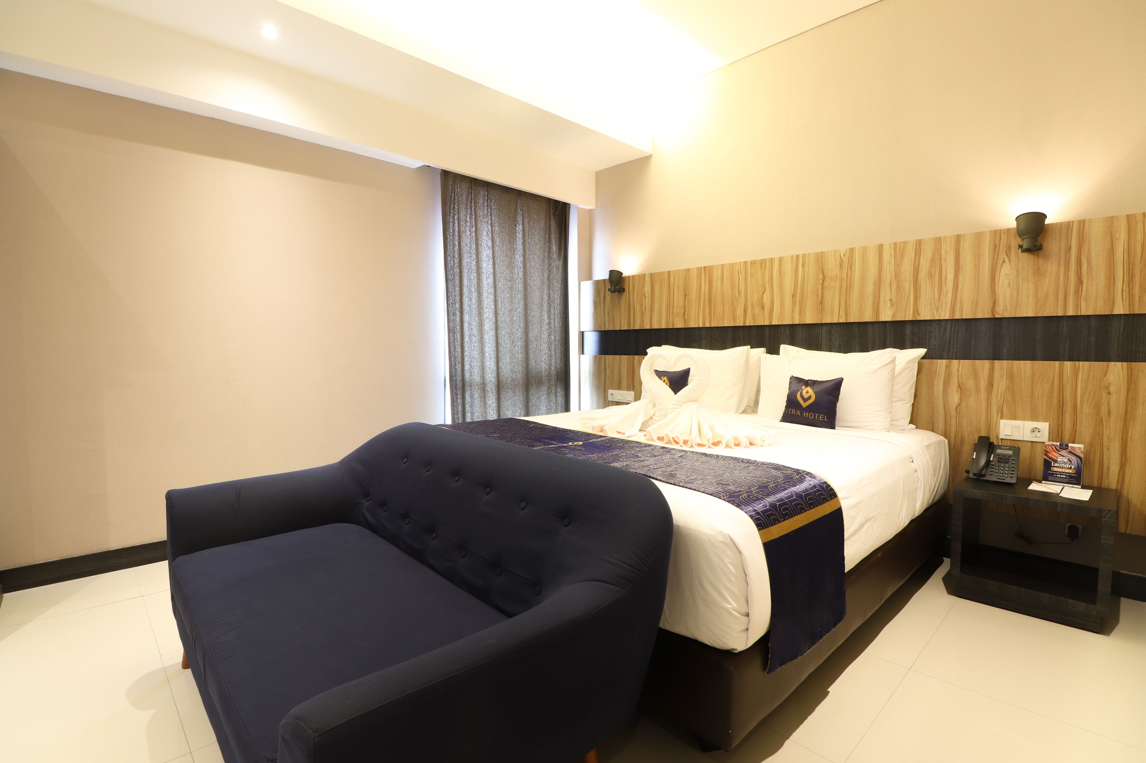 Une chambre d’hôtel avec un lit, un fauteuil bleu foncé, une tête de lit en bois, une table de chevet avec un téléphone et une lampe, et une fenêtre avec des rideaux.