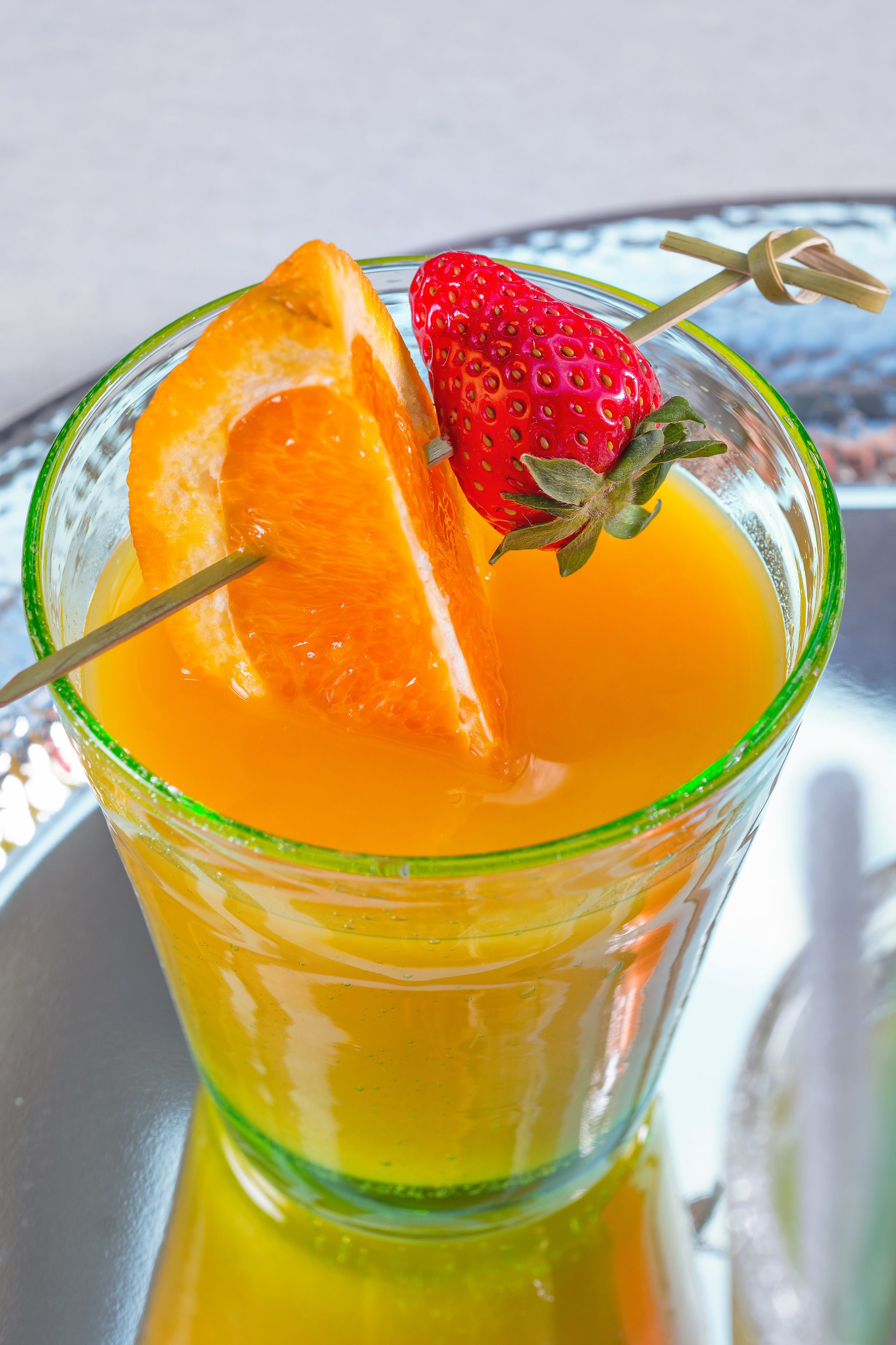A glass with a slice of orange and a strawberry on a reflective surface.