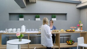 A modern outdoor dining area with a long table, chairs, and a counter with food and drinks. There are potted plants on the counter and a white table with a vase of flowers. - Lake Spirit Boutique Hotel & Spa (Ioannina)