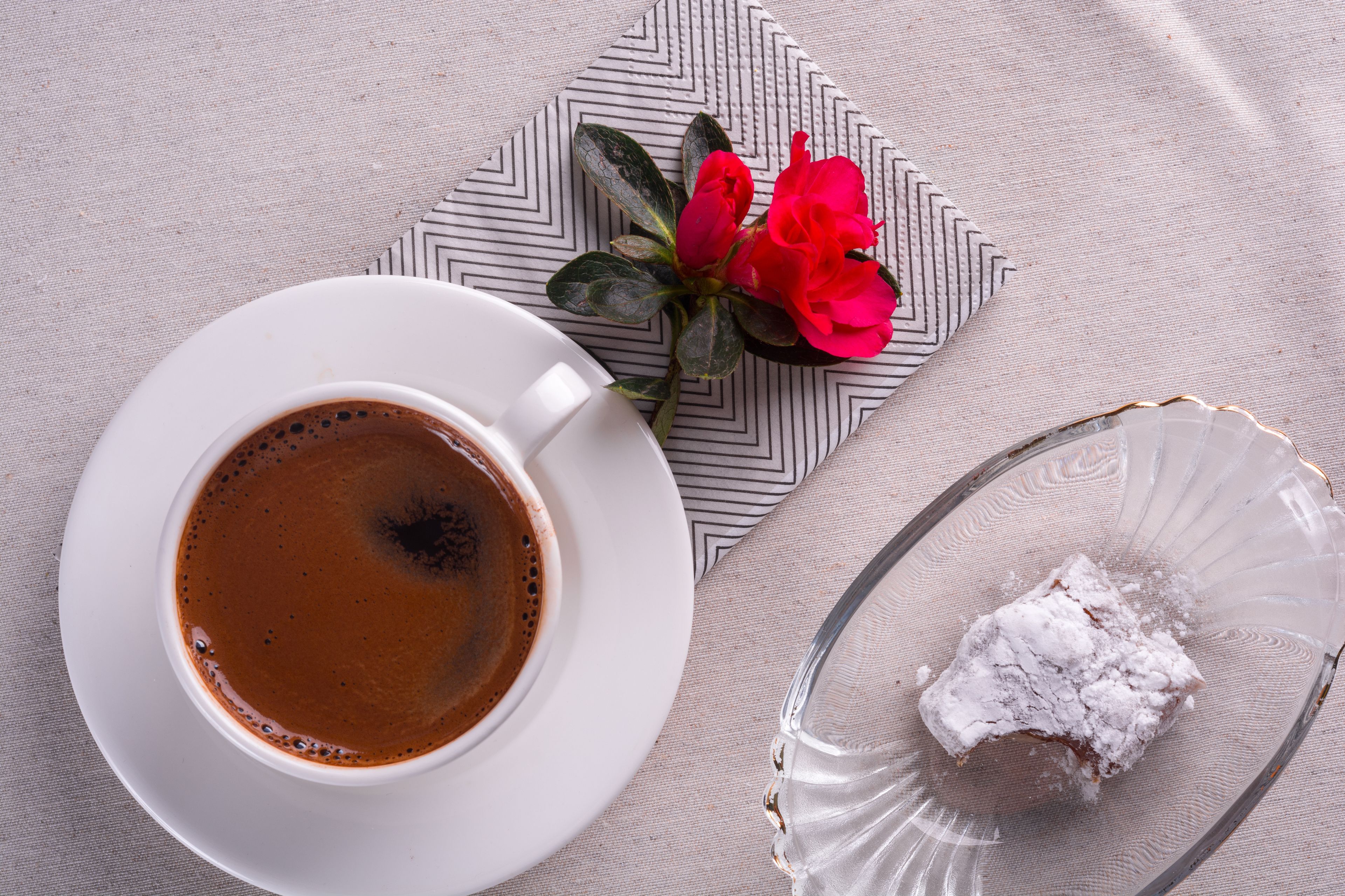 Una taza de café blanca sobre un platito, un plato de vidrio con un postre y una flor roja sobre una servilleta estampada.