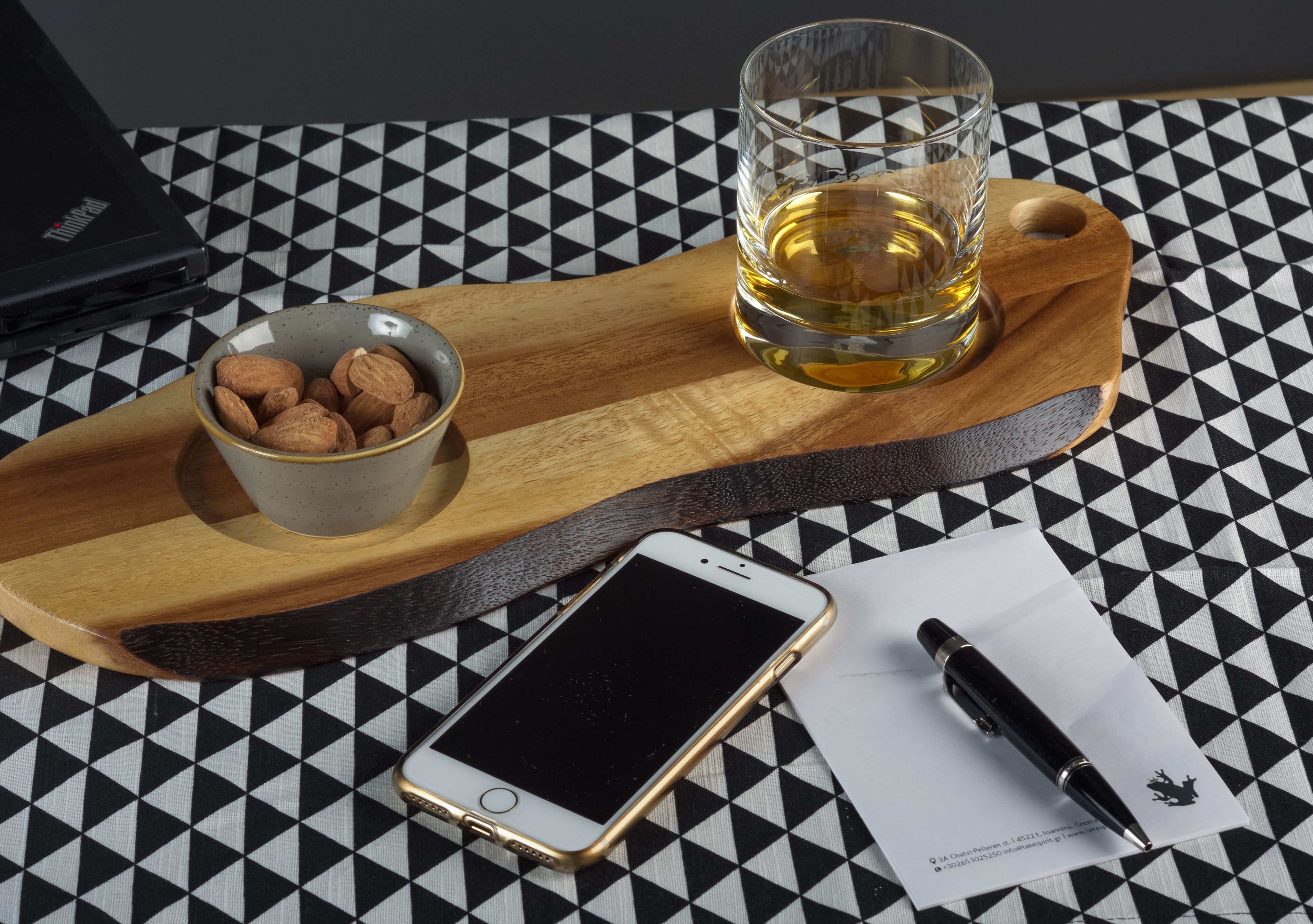 Un teléfono inteligente, una copa de whisky, un cuenco de almendras, un bolígrafo y un ordenador portátil sobre un mantel a cuadros.