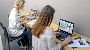 Two individuals seated at a wooden table with laptops, one of which displays a travel website.