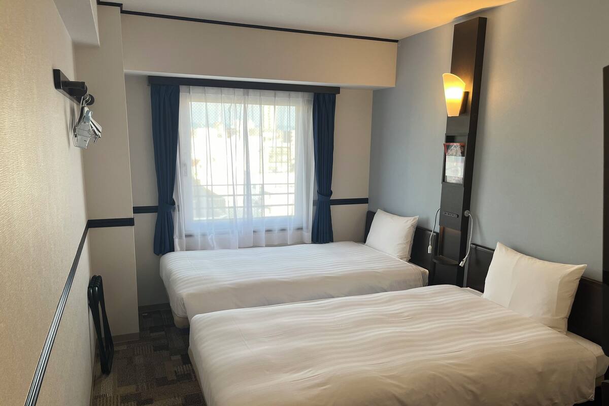 Une chambre d’hôtel avec deux lits, une grande fenêtre avec des rideaux légers, un téléphone fixé au mur et une table de chevet avec une lampe.
