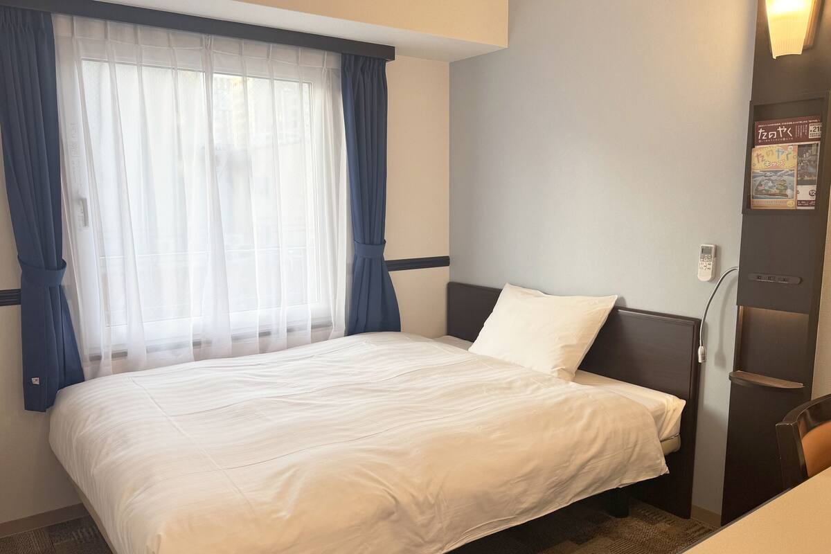 Une chambre d’hôtel comprenant un lit, un bureau, une chaise, une fenêtre avec des rideaux, un téléphone fixé au mur et un tableau d’affichage avec des documents.