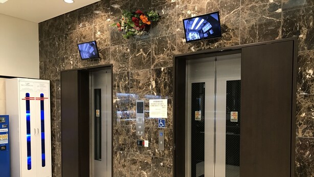 An elevator area with marble walls, two elevators, a cinema machine, and a flower arrangement. - Toyoko Inn Osaka Hommachi No.2 (Osaka)