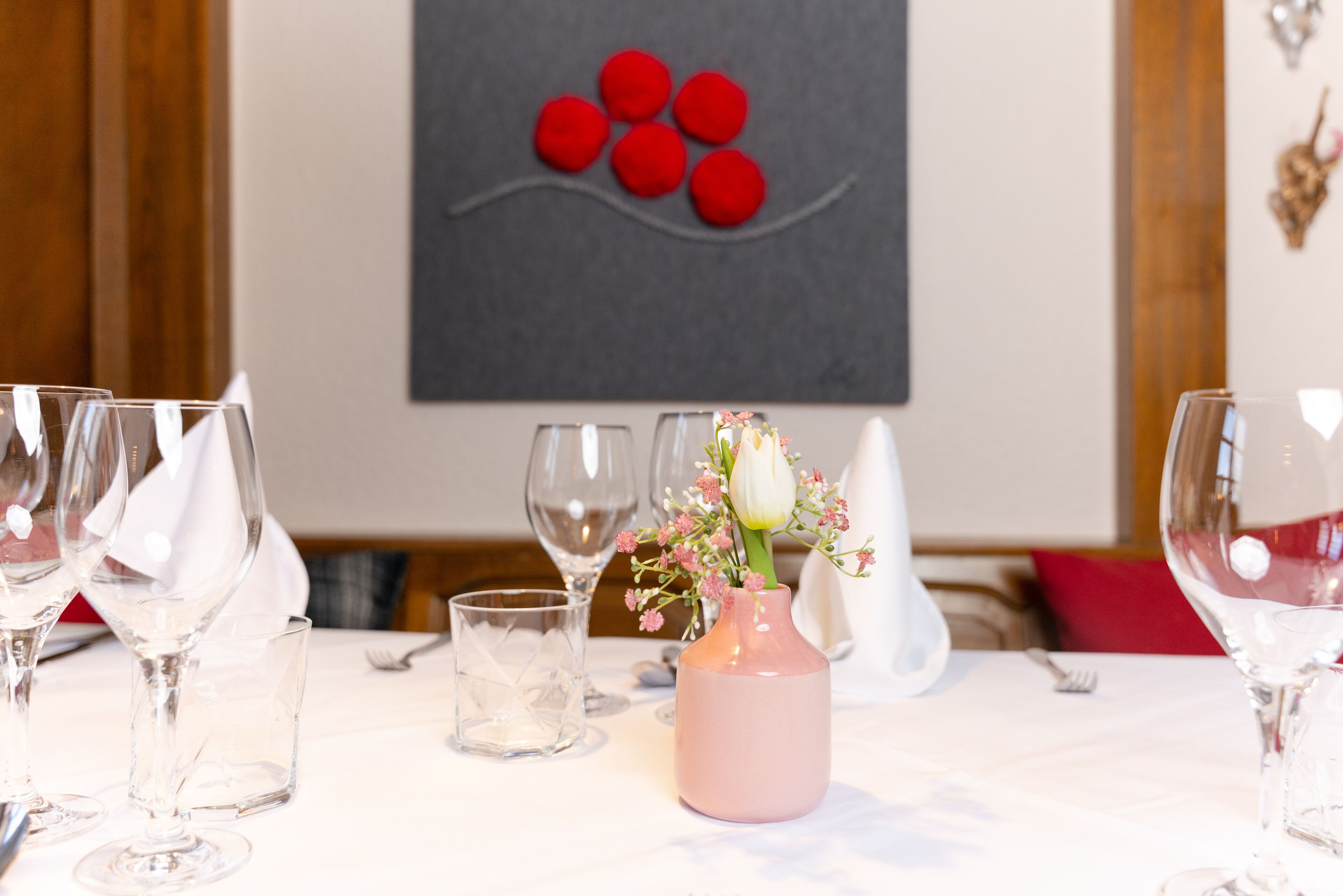 A dining table with empty wine glasses, a pink vase with flowers, and a decorative wall piece.