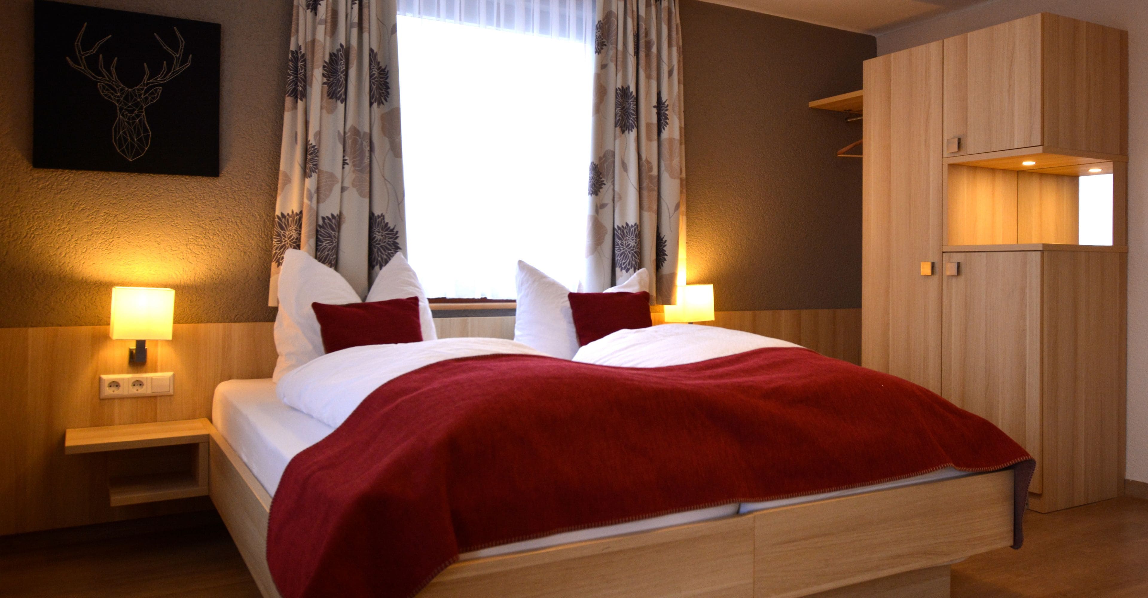 Quarto de hotel com cama, duas luminárias de cabeceira, janela com cortinas, uma cabeça de veado como objeto de arte na parede e um guarda-roupa de madeira.