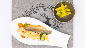 A white plate with a piece of fish, lemon wedges, and garnishes on a gray placemat with a deer and leaf design.