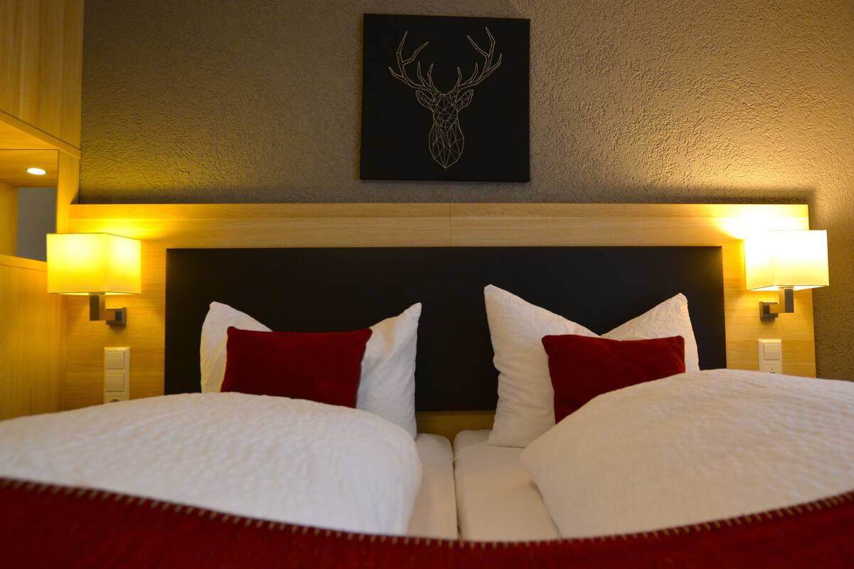 A double bed with white and red bedding, two wall-mounted lamps, and a deer head artwork above the bed.