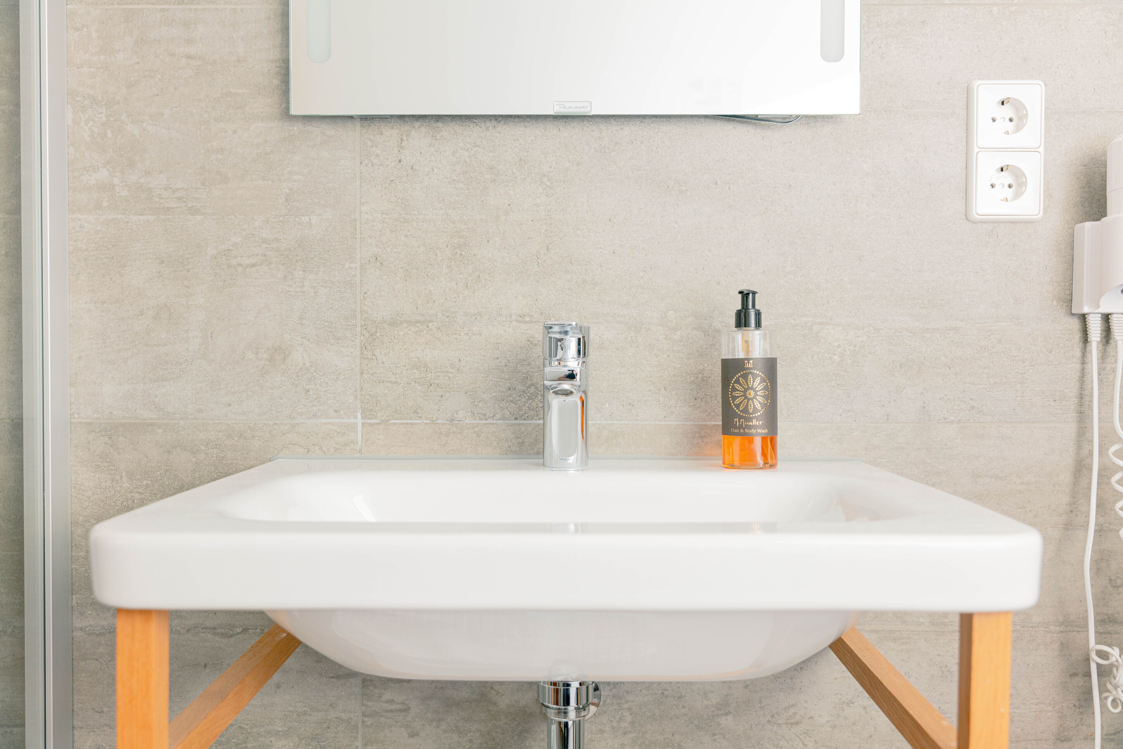 Un lavabo blanc dans une salle de bain, avec un robinet chromé, un flacon de savon et un miroir au-dessus.
