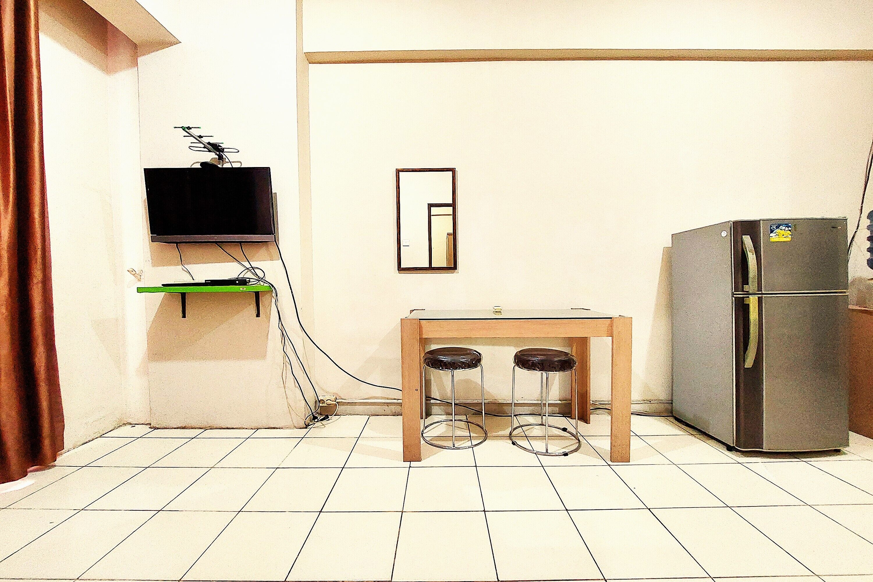 Una pequeña cocina americana con un televisor montado en la pared, un refrigerador y una mesa de madera con dos taburetes.