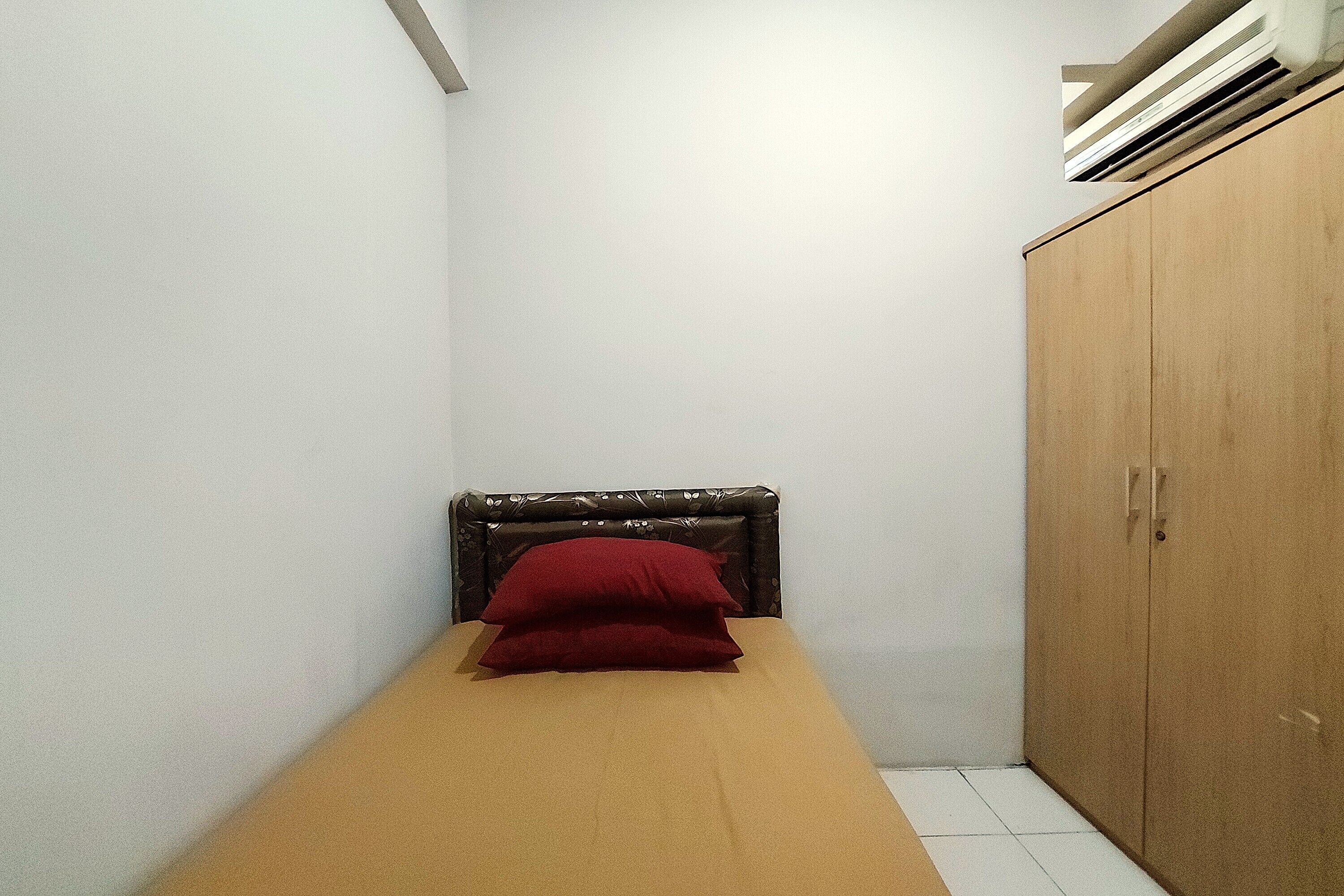 A small, single-bed room with a wooden headboard and a red pillow. There is a wooden wardrobe to the right.