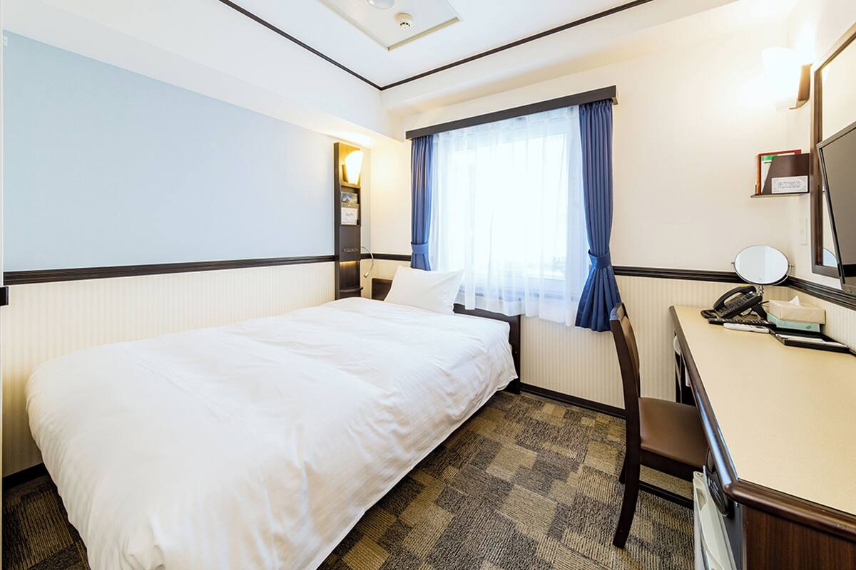 A hotel room with a bed, a desk, a chair, a window with curtains, a bedside lamp, a mirror, and a telephone.