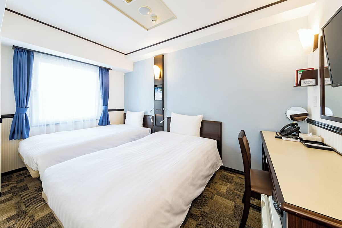 A hotel room with two beds, a desk with a telephone, a mirror, and a window with curtains.
