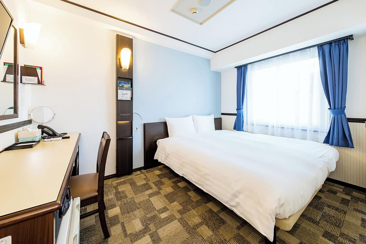 A hotel room with a bed, a desk with a telephone, a chair, a mirror, and a window with curtains.