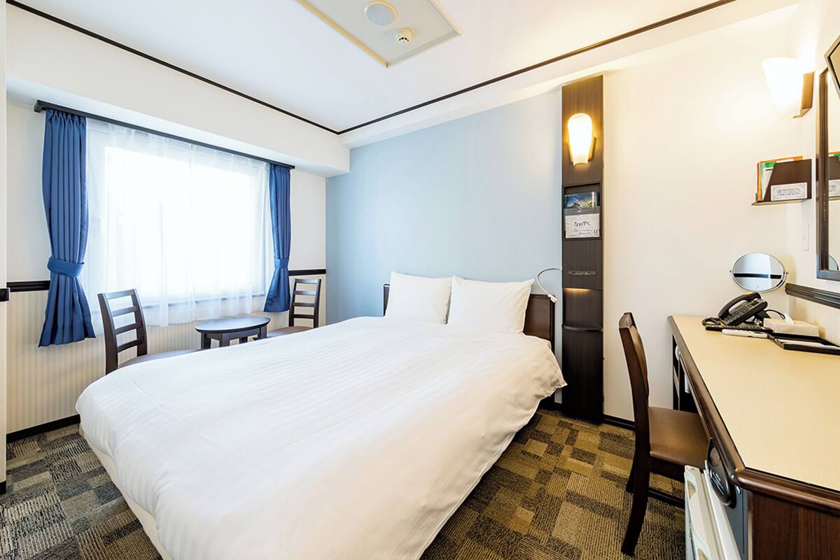 A hotel room with a large bed, a desk with a telephone, a chair, a window with curtains, and a wall-mounted light.