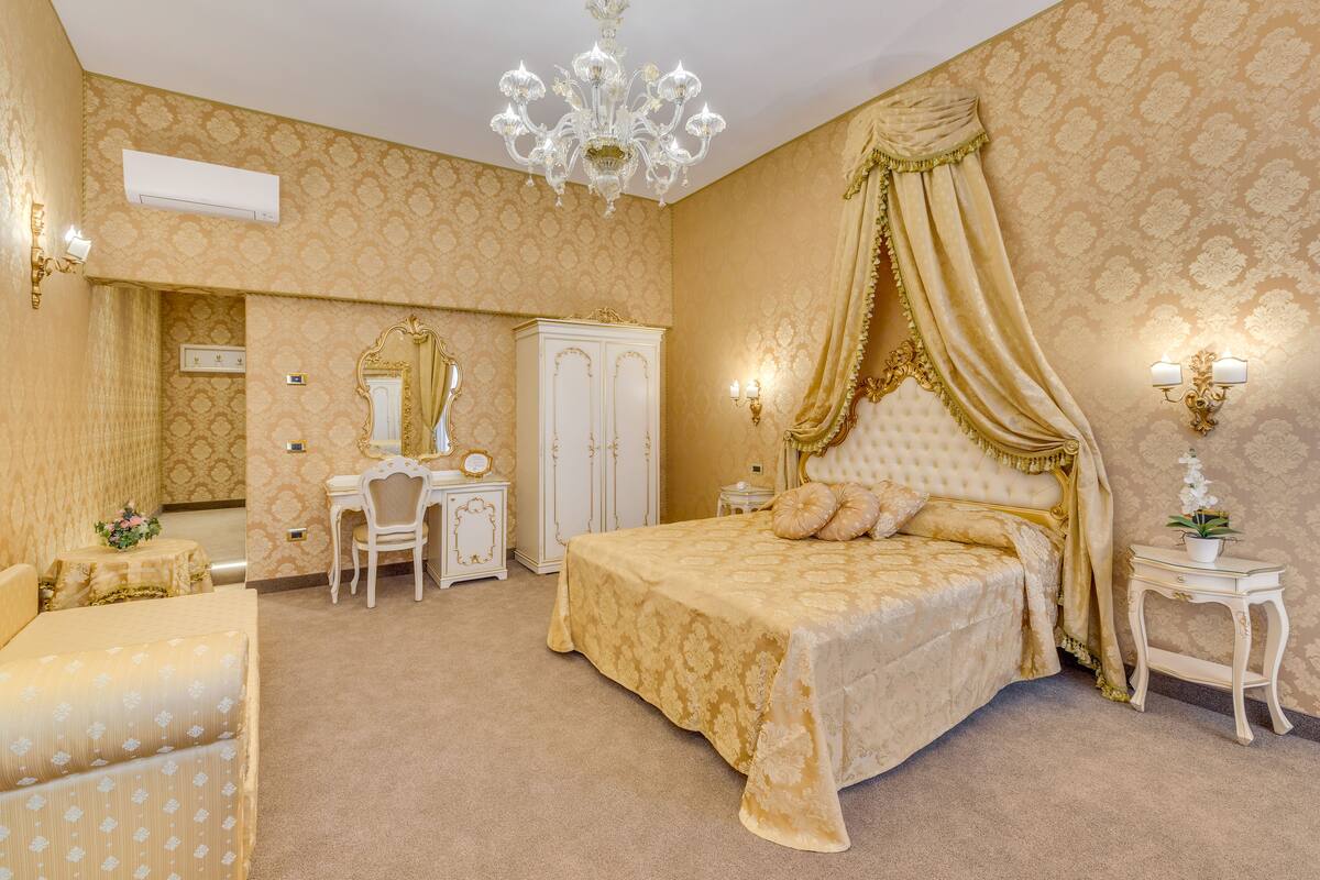 A luxurious bedroom with a large bed, a vanity with a mirror, a dressing table, and a chandelier.