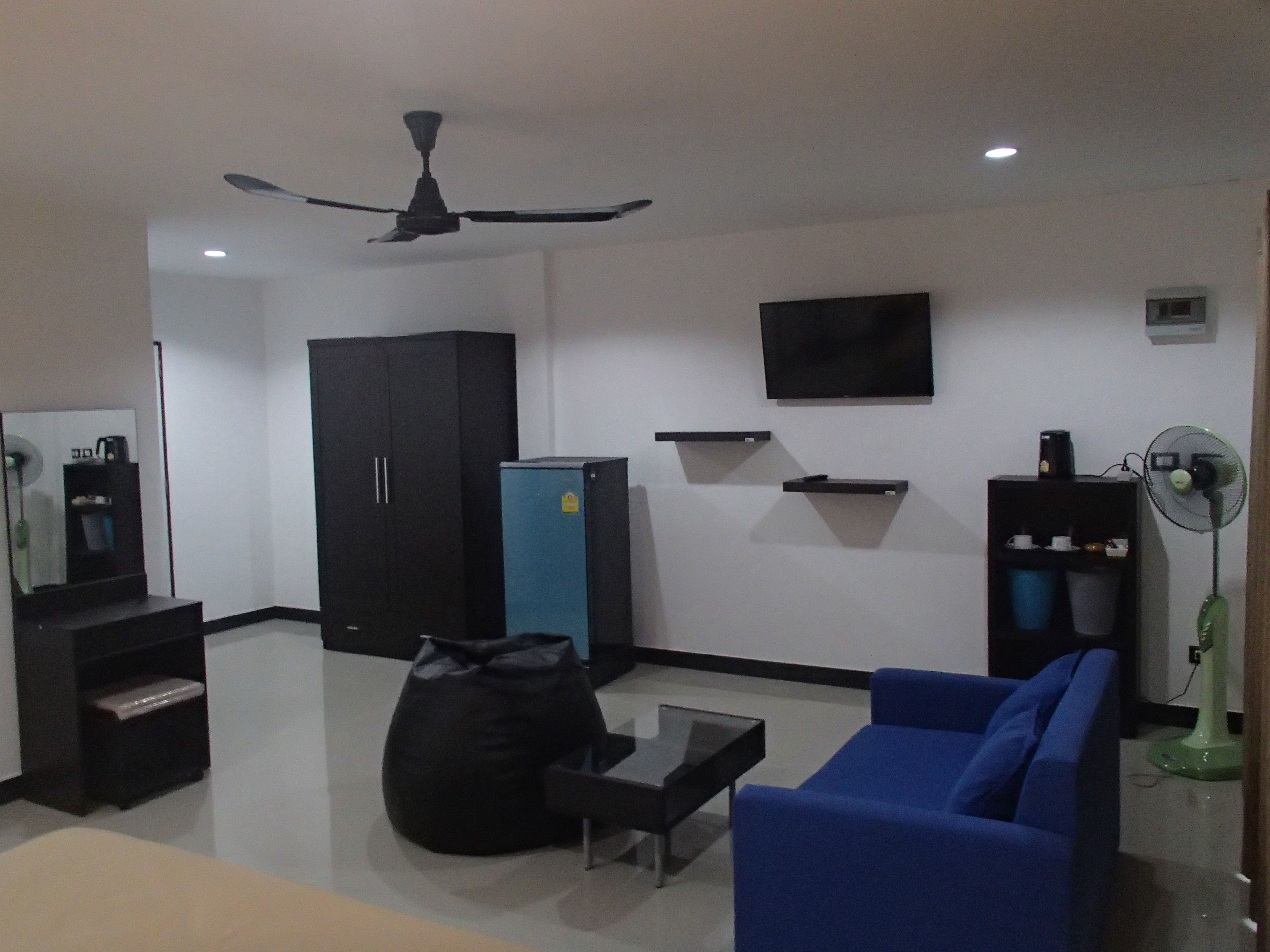 Una sala de estar con ventilador de techo, un sofá azul, un sillón de peluche negro, una mesita, un televisor y un refrigerador.