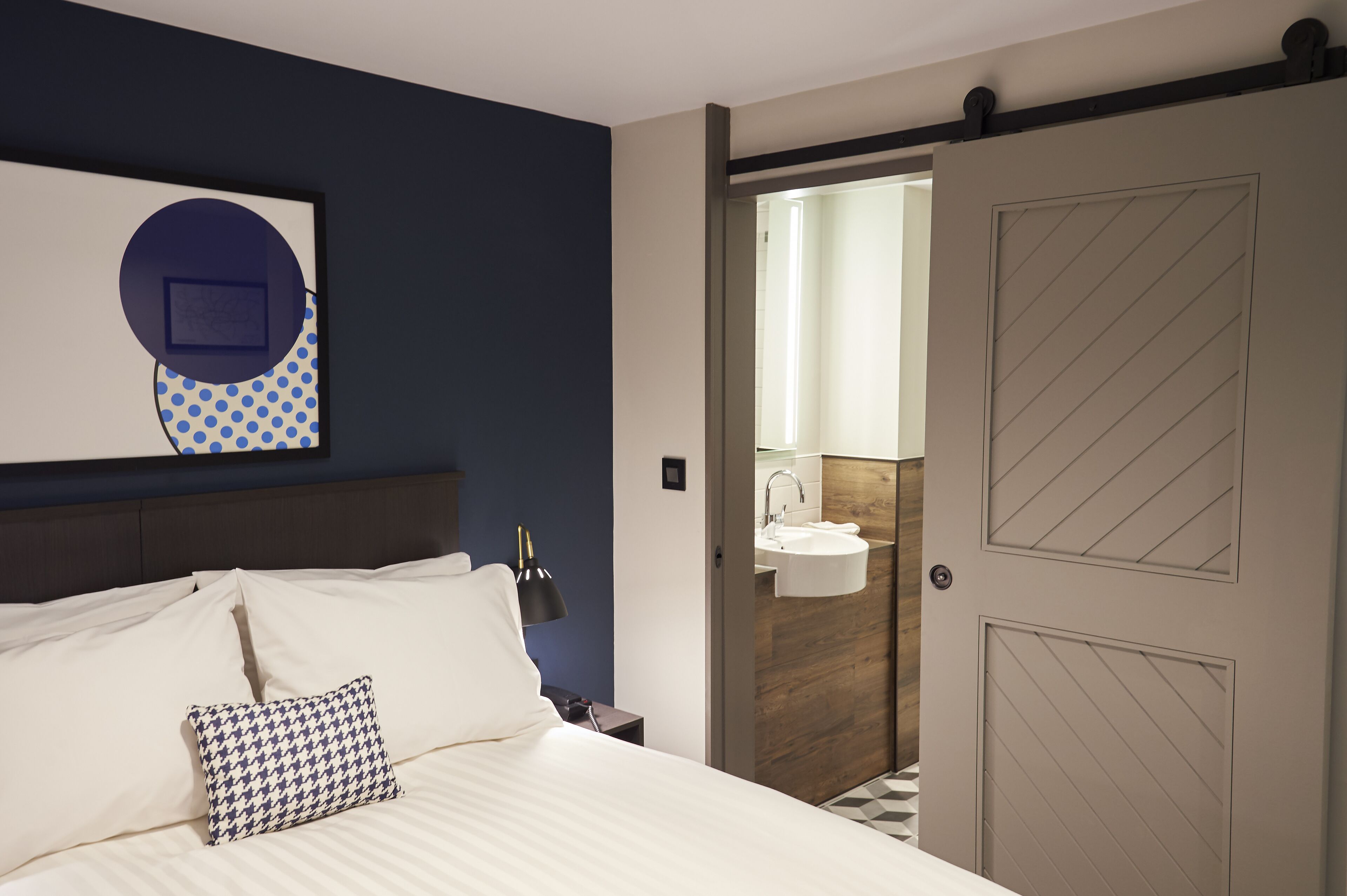A hotel room with a bed, a nightstand, a lamp, a wall art piece, a door leading to a bathroom with a sink, and a patterned pillow.