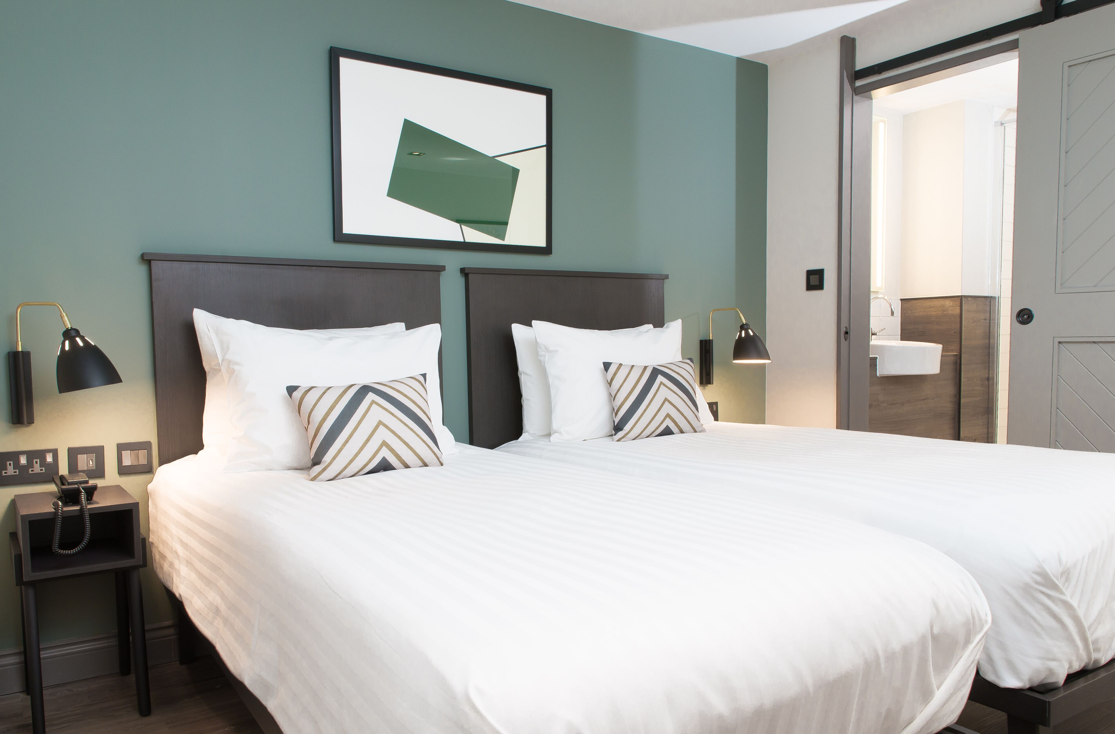 A double bed with white linens and striped pillows, a bedside table with a lamp, and a framed picture on the wall.