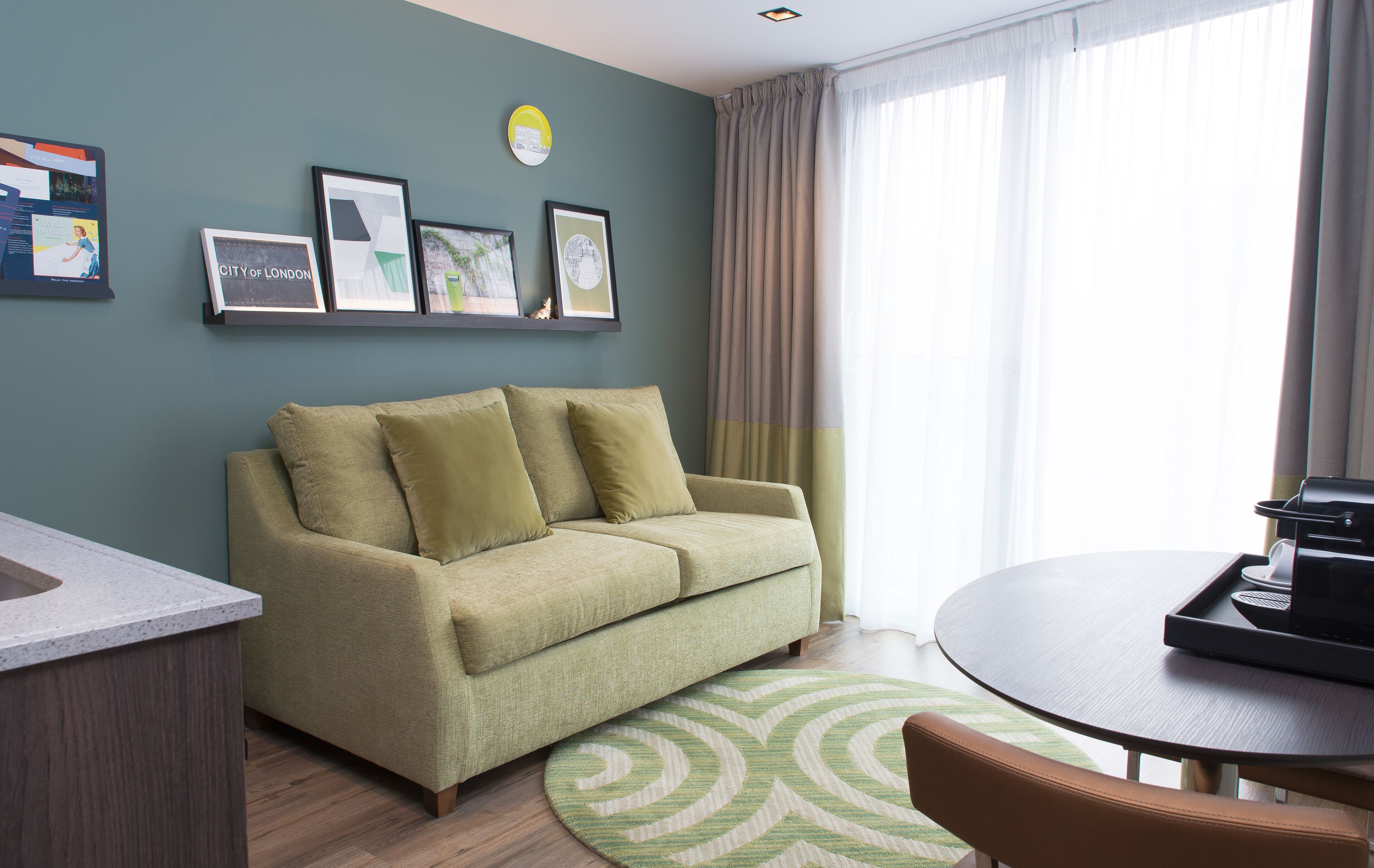 A modern living room with a sofa, a round table, and a coffee machine. There are framed pictures on the wall and a window with curtains.