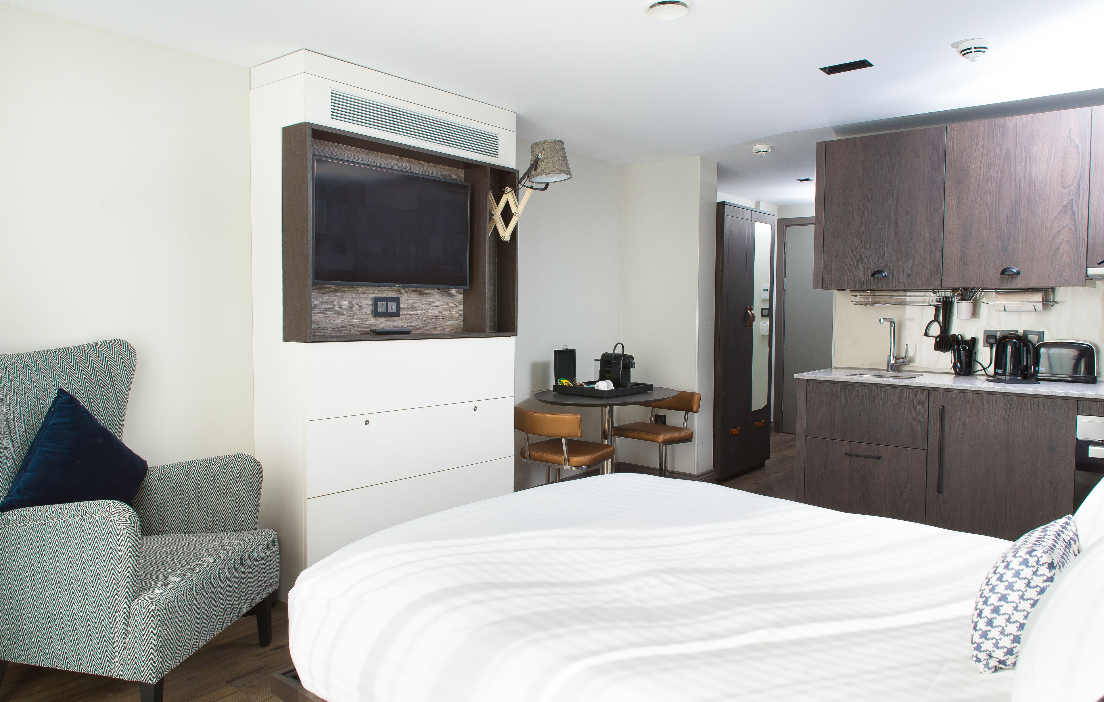 Une chambre d’hôtel comprenant un lit, un téléviseur fixé au mur, une petite table à manger avec des chaises et un coin cuisine.