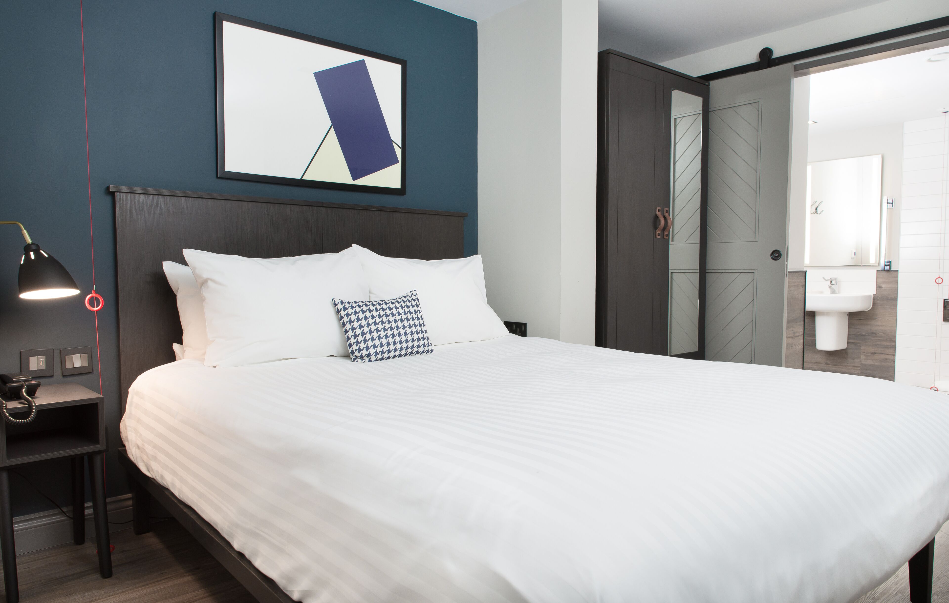 A neatly made bed with white bedding and a patterned pillow, a bedside table with a lamp, and a framed picture on the wall.