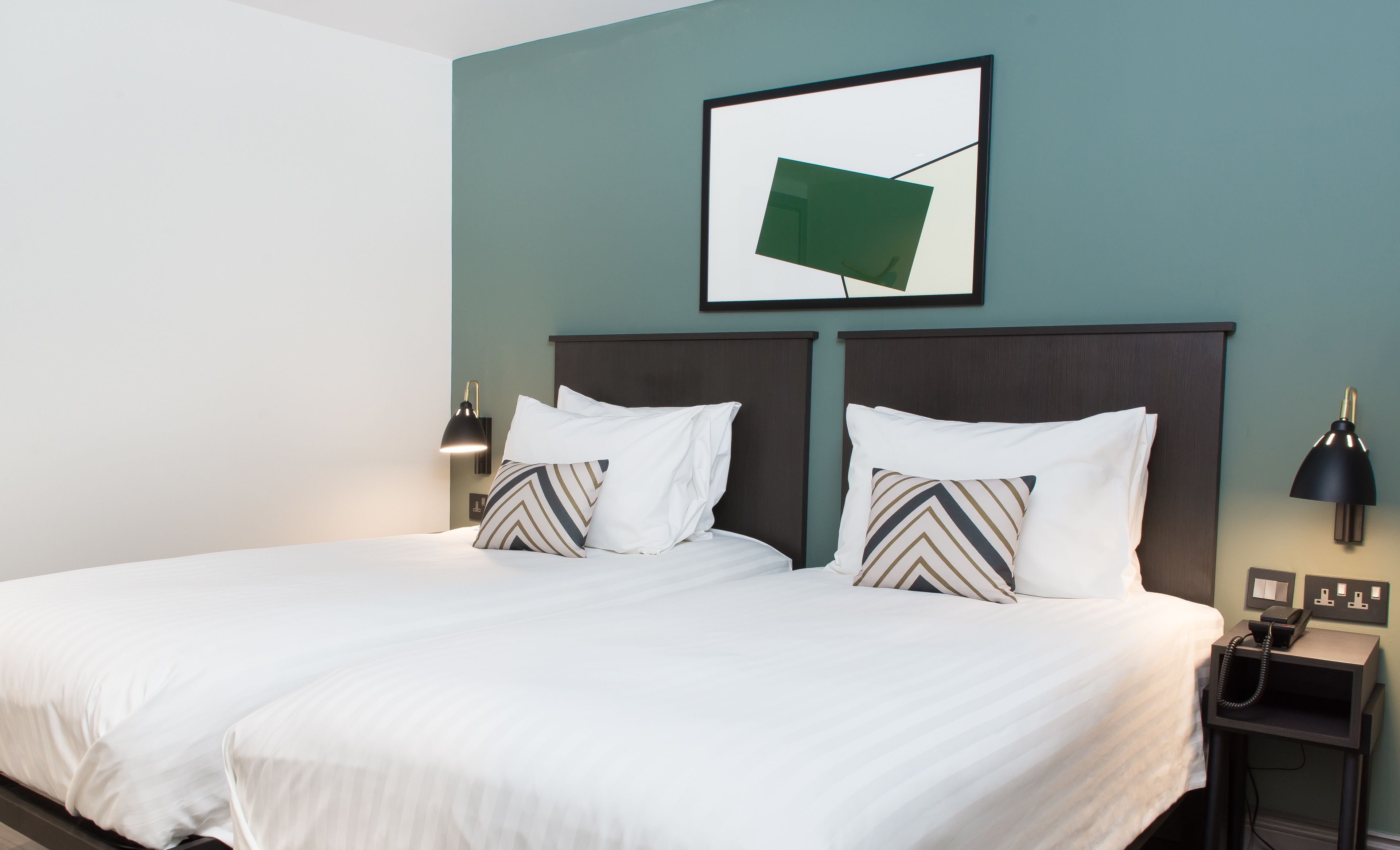 A double bed with white linens and two pillows, a bedside table with a lamp, and a framed picture on the wall.