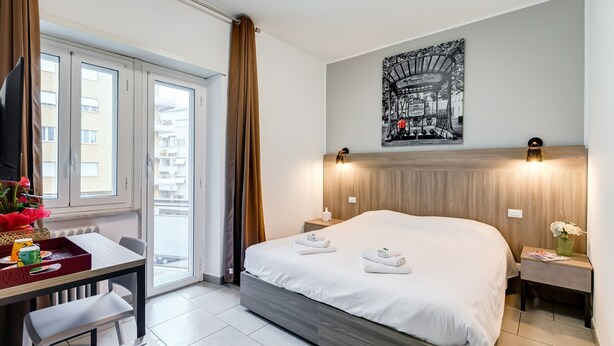 A modern hotel room with a bed, a desk with a tray, a chair, a small table, and a nightstand with a lamp. - B&B Casa Der Poeta (Rome)