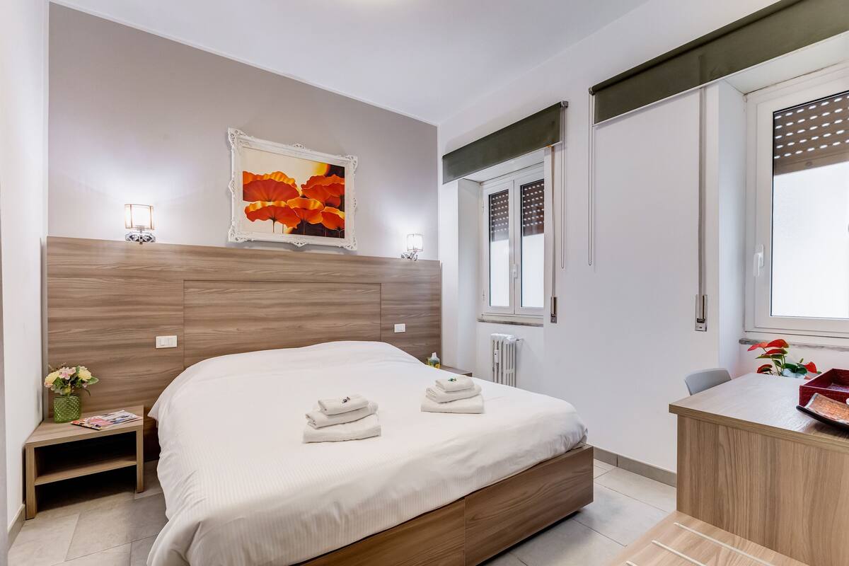 A modern hotel room with a wooden headboard, a large bed, a small bedside table with a lamp, a desk with a chair, and a window with blinds.