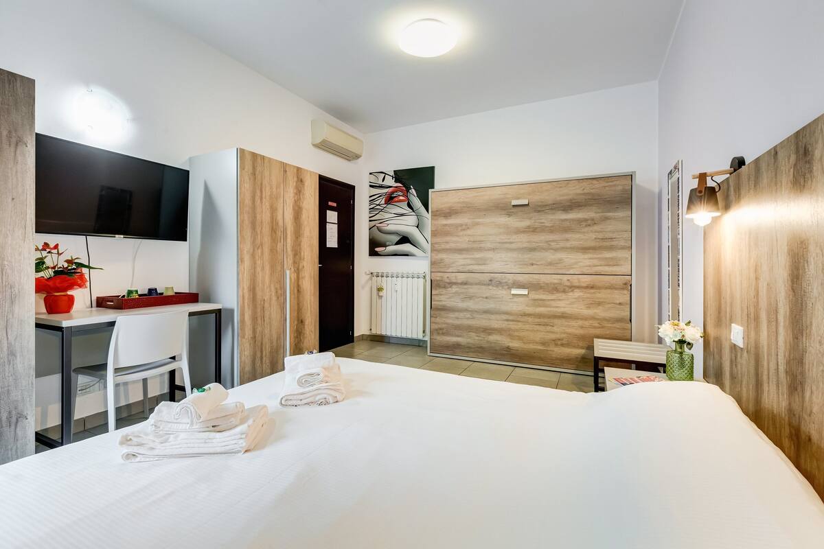 A modern hotel room with a large bed, a desk with a chair, a flat-screen TV, and a wooden wardrobe.