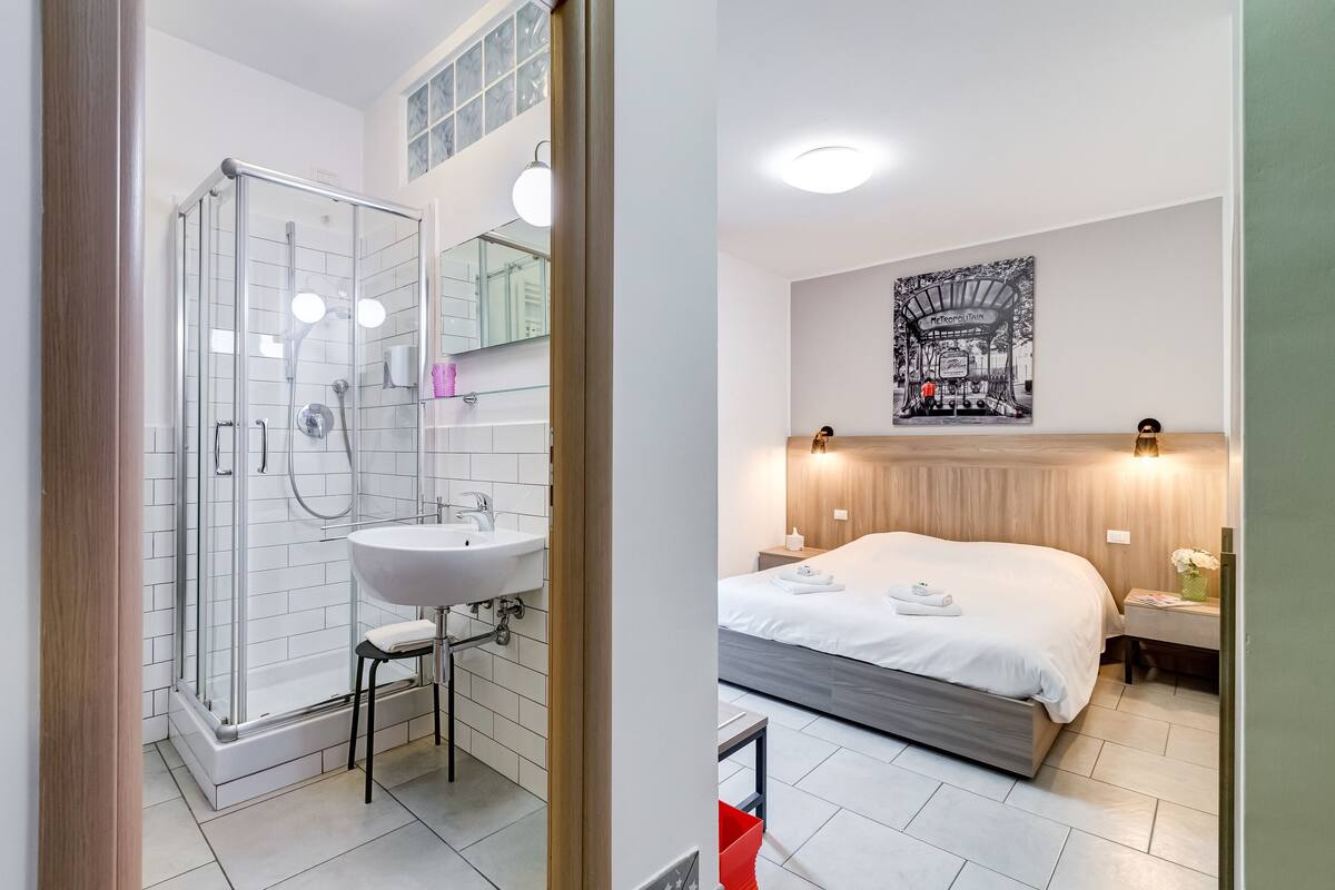A modern bathroom with a glass shower enclosure, a sink, and a mirror. The room features a large bed with a wooden headboard, a bedside table with a vase of flowers, and a wall-mounted artwork.