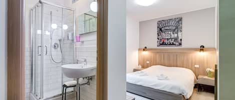 Une salle de bain moderne avec une douche à parois de verre, un lavabo et un miroir. La pièce est dotée d’un grand lit avec une tête de lit en bois, d’une table de chevet ornée d’un vase de fleurs et d’une œuvre d’art accrochée au mur.