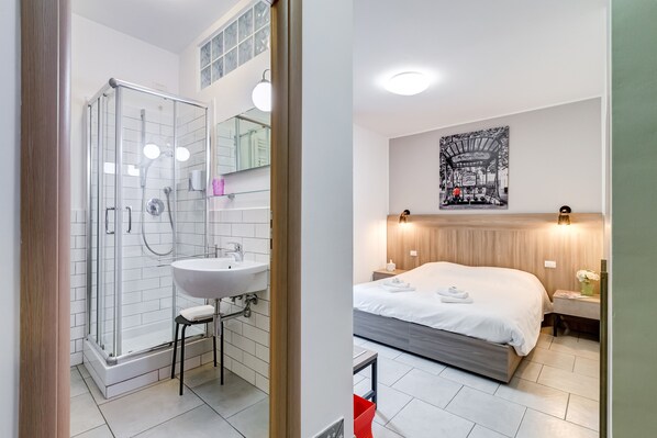 A modern bathroom with a glass shower enclosure, a sink, and a mirror. The room features a large bed with a wooden headboard, a bedside table with a vase of flowers, and a wall-mounted artwork. - B&B Casa Der Poeta (Rome)