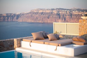 A sunlit outdoor seating area with cushions and a pool, overlooking a cliff and sea.