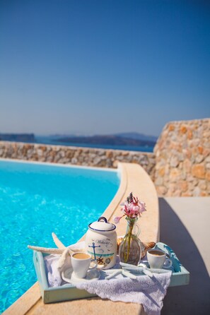 A tray with a teapot, cups, and a vase of flowers by a pool with a stone wall and a view of the sea.