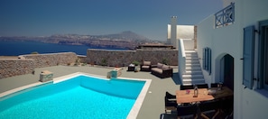A pool area with a view of the sea and a mountain, stone walls, and outdoor furniture.