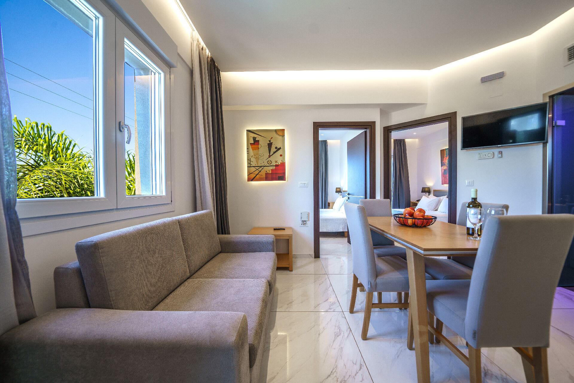 A modern hotel room with a sofa, dining table, chairs, a TV, and a view of greenery outside.