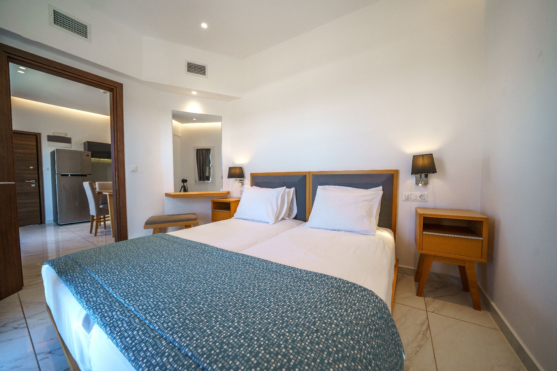 A hotel room with a large bed, bedside tables, a mirror, and a kitchenette in the background.