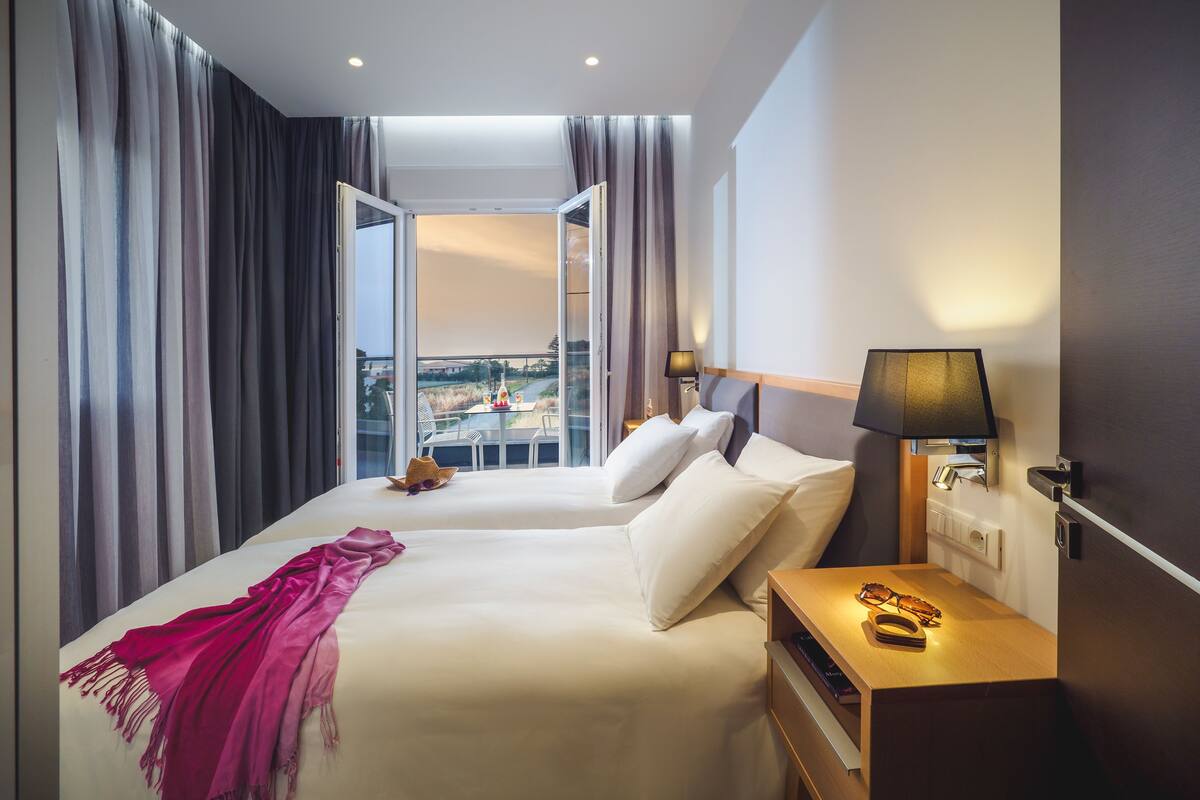 A hotel room with two beds, a balcony, and a view of the cityscape.