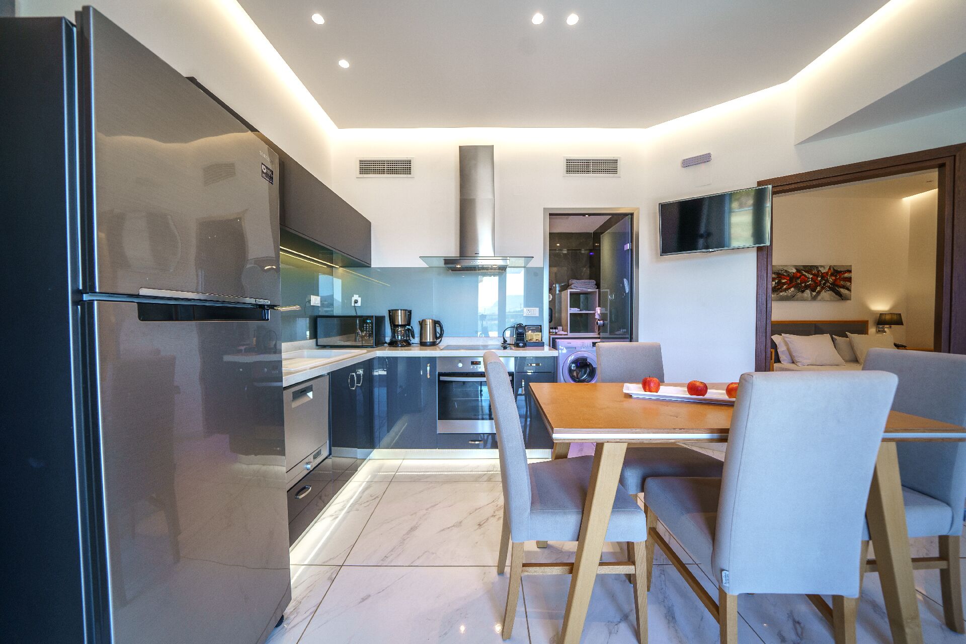 A modern kitchen with a refrigerator, microwave, and coffee maker. There is a dining table with chairs and a TV mounted on the wall.