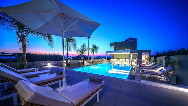 Un área de piscina con sillones de descanso y sombrillas, un edificio moderno al fondo y vistas al atardecer.