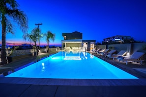 A swimming pool with lounge chairs, a covered seating area, and a view of the city at dusk. - Elysian Suites (Hersonissos)