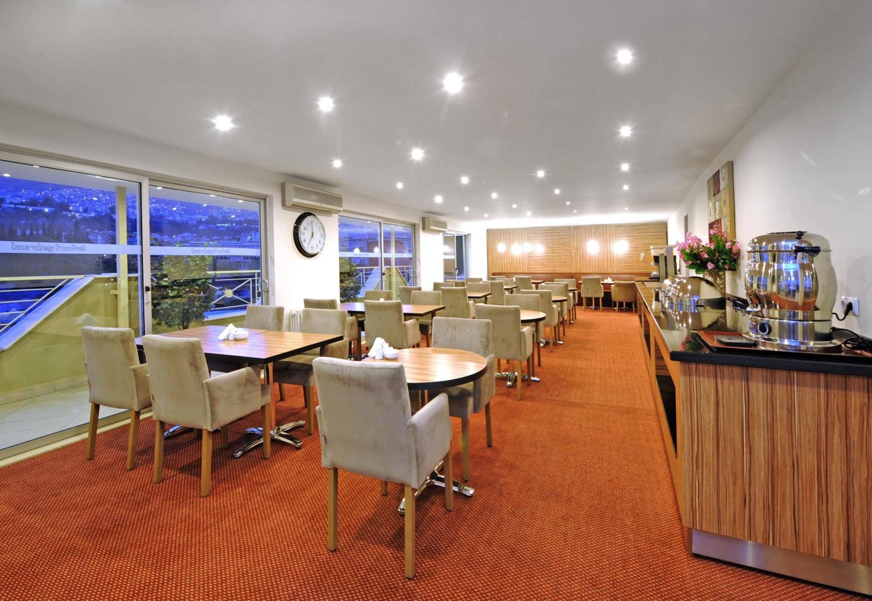 Un comedor de hotel con mesas y sillas, un reloj en la pared y vistas de la ciudad a través de grandes ventanales.