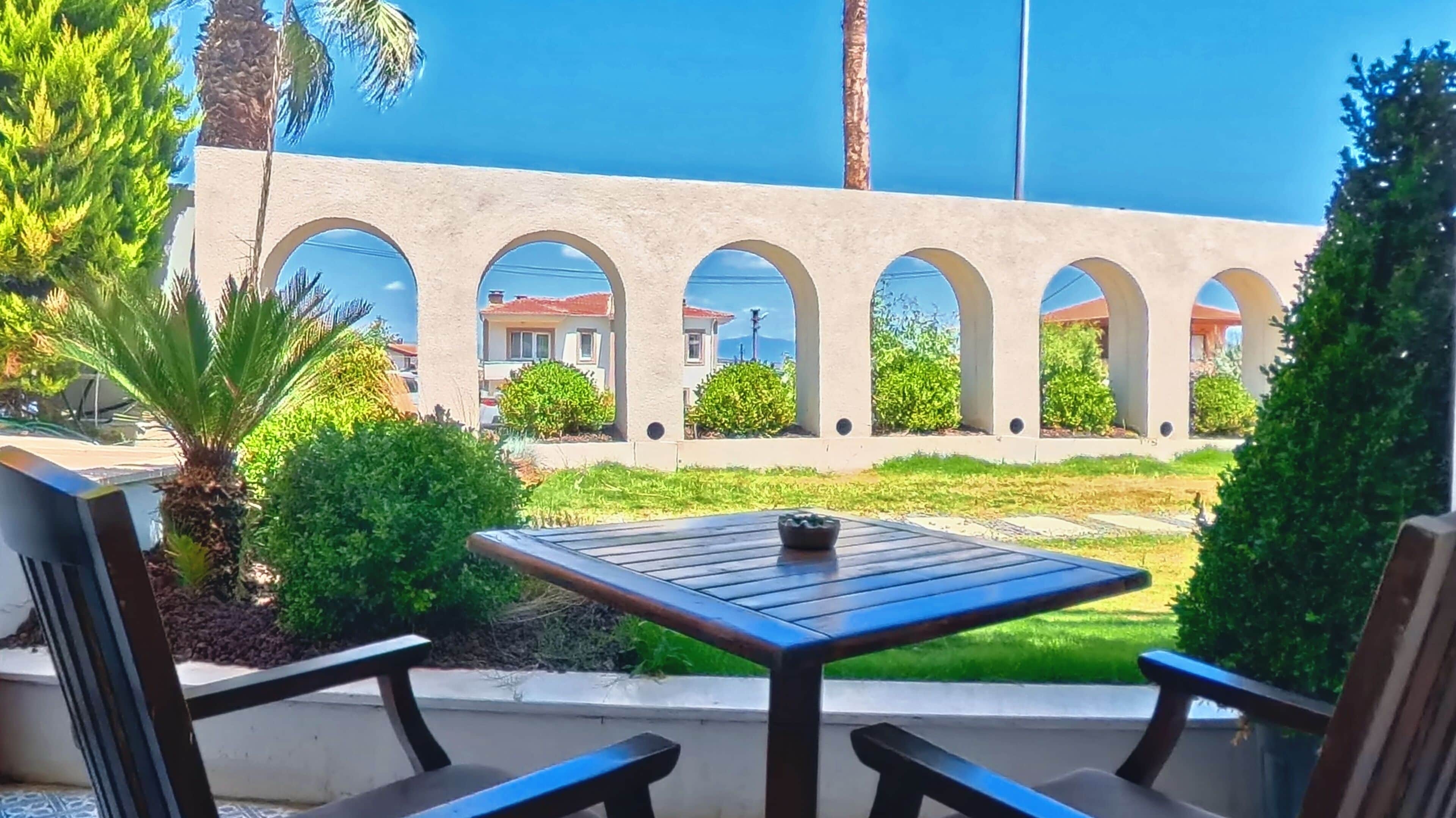 An outdoor patio with a table and chairs, surrounded by greenery and a view of buildings.